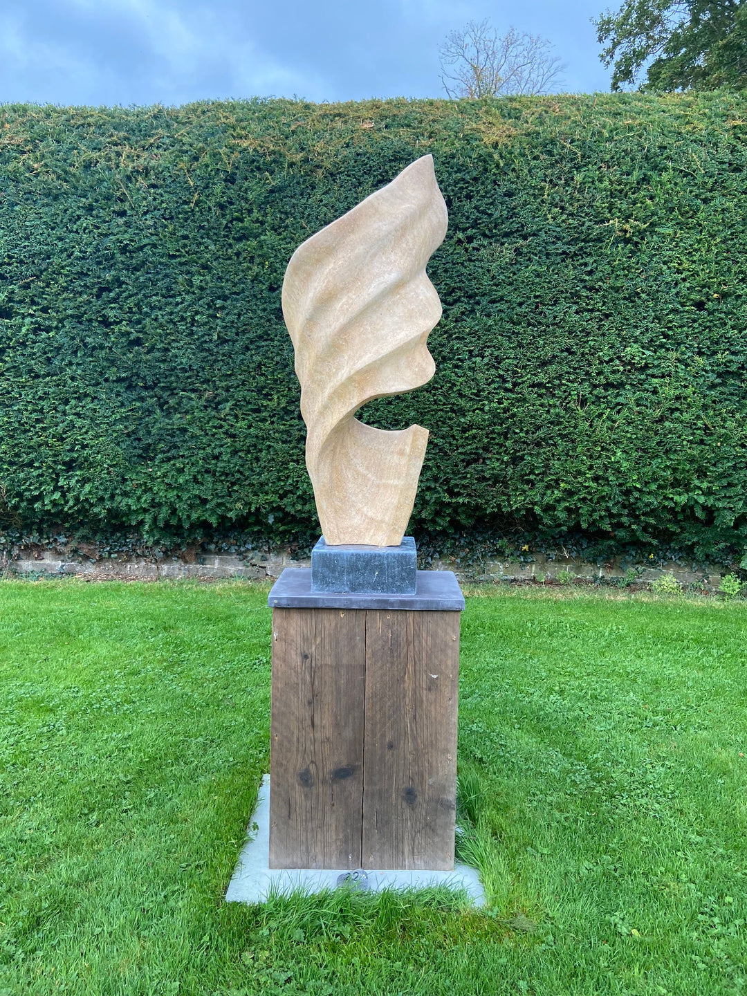 Billow 1 by Brendan Cullen, carved from Ancaster Weatherbed Limestone, a tall sandstone-toned sculpture with soft flowing ridges that rise in a twisting, wave-like form, mounted on a dark stone base.