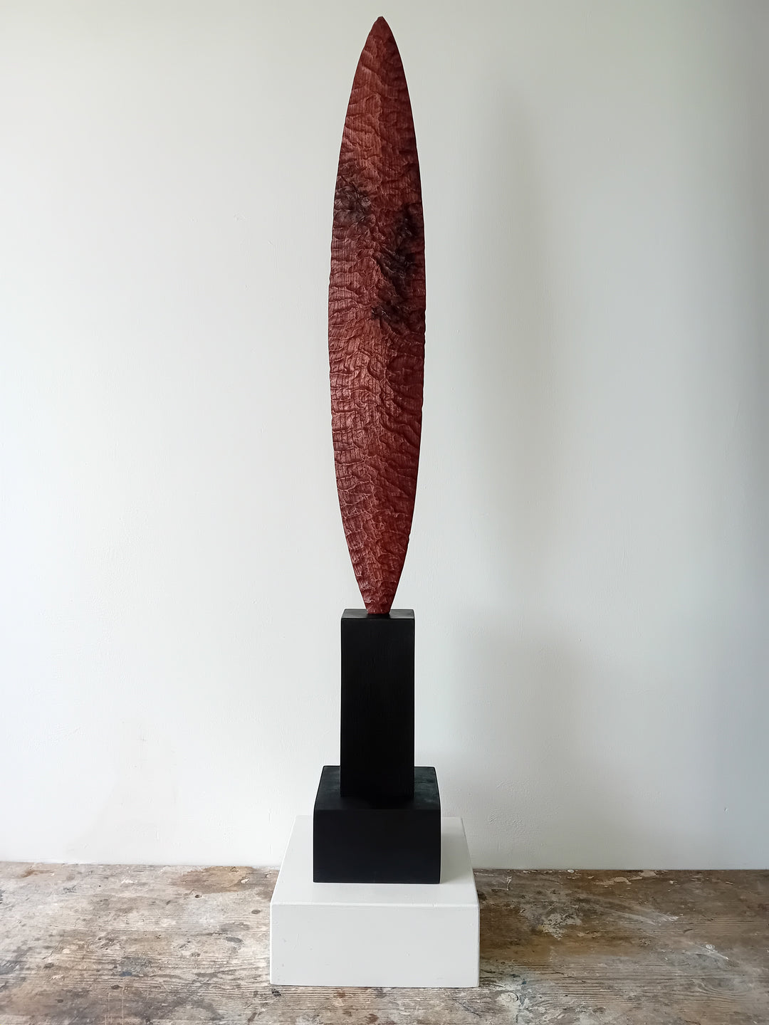 Blade by Nicola Beattie – a tall, slender, hand-carved elm sculpture with an elongated leaf-like or blade-shaped form. The surface is deeply textured with rhythmic chisel marks, finished in rich reddish-brown tones with areas of darker burnished grain and natural patterning. Mounted on a tall black rectangular base atop a square plinth, the piece stands vertically like a minimalist totem. Its organic contours and refined symmetry evoke ancient tools or ceremonial artefacts, blending contemporary abstraction