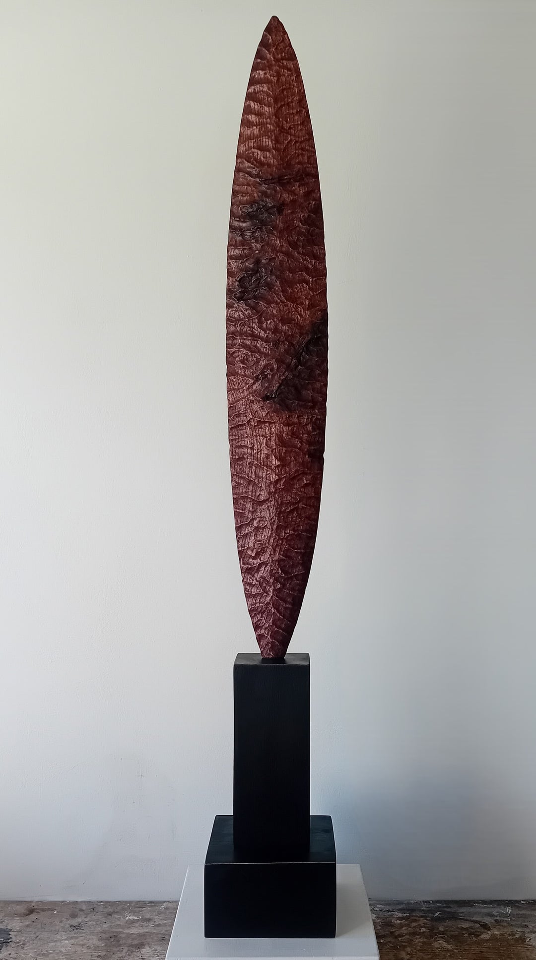 Blade by Nicola Beattie – a tall, slender, hand-carved elm sculpture with an elongated leaf-like or blade-shaped form. The surface is deeply textured with rhythmic chisel marks, finished in rich reddish-brown tones with areas of darker burnished grain and natural patterning. Mounted on a tall black rectangular base atop a square plinth, the piece stands vertically like a minimalist totem. Its organic contours and refined symmetry evoke ancient tools or ceremonial artefacts, blending contemporary abstraction