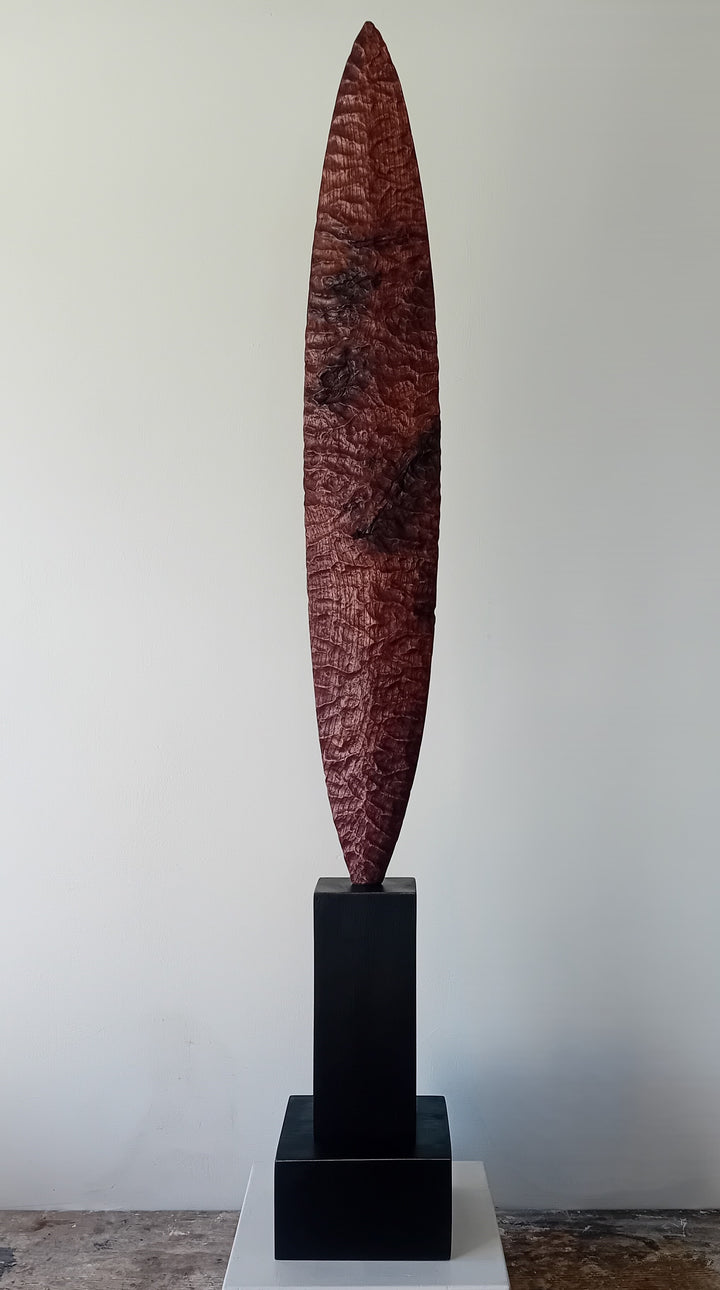 Blade by Nicola Beattie – a tall, slender, hand-carved elm sculpture with an elongated leaf-like or blade-shaped form. The surface is deeply textured with rhythmic chisel marks, finished in rich reddish-brown tones with areas of darker burnished grain and natural patterning. Mounted on a tall black rectangular base atop a square plinth, the piece stands vertically like a minimalist totem. Its organic contours and refined symmetry evoke ancient tools or ceremonial artefacts, blending contemporary abstraction