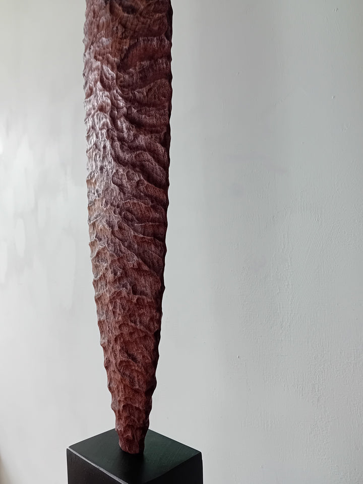Blade by Nicola Beattie – a tall, slender, hand-carved elm sculpture with an elongated leaf-like or blade-shaped form. The surface is deeply textured with rhythmic chisel marks, finished in rich reddish-brown tones with areas of darker burnished grain and natural patterning. Mounted on a tall black rectangular base atop a square plinth, the piece stands vertically like a minimalist totem. Its organic contours and refined symmetry evoke ancient tools or ceremonial artefacts, blending contemporary abstraction