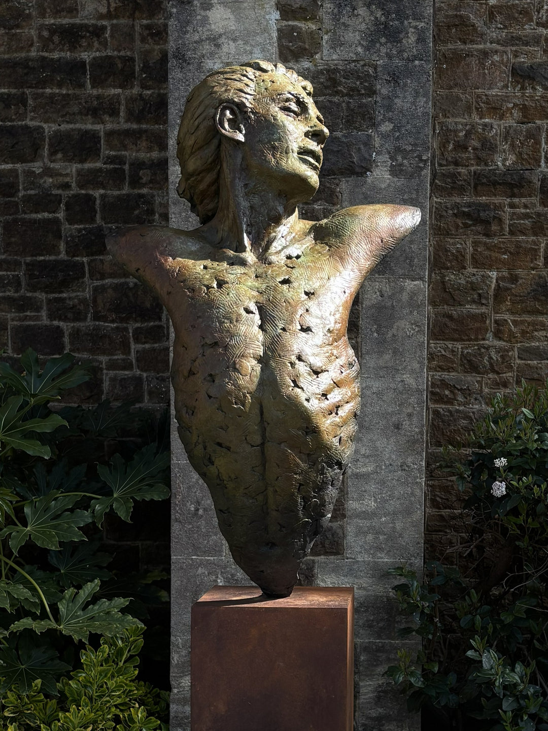Nicola Godden Bliss Icarus bronze sculpture portraying a stylised human figure rising upward with arms extended and head turned, evoking a moment of release and quiet strength. The elongated form suggests lift and motion while remaining grounded through a solid Corten steel base. The textured bronze surface carries depth and variation, with light catching across its contours to enhance the sense of movement and presence.