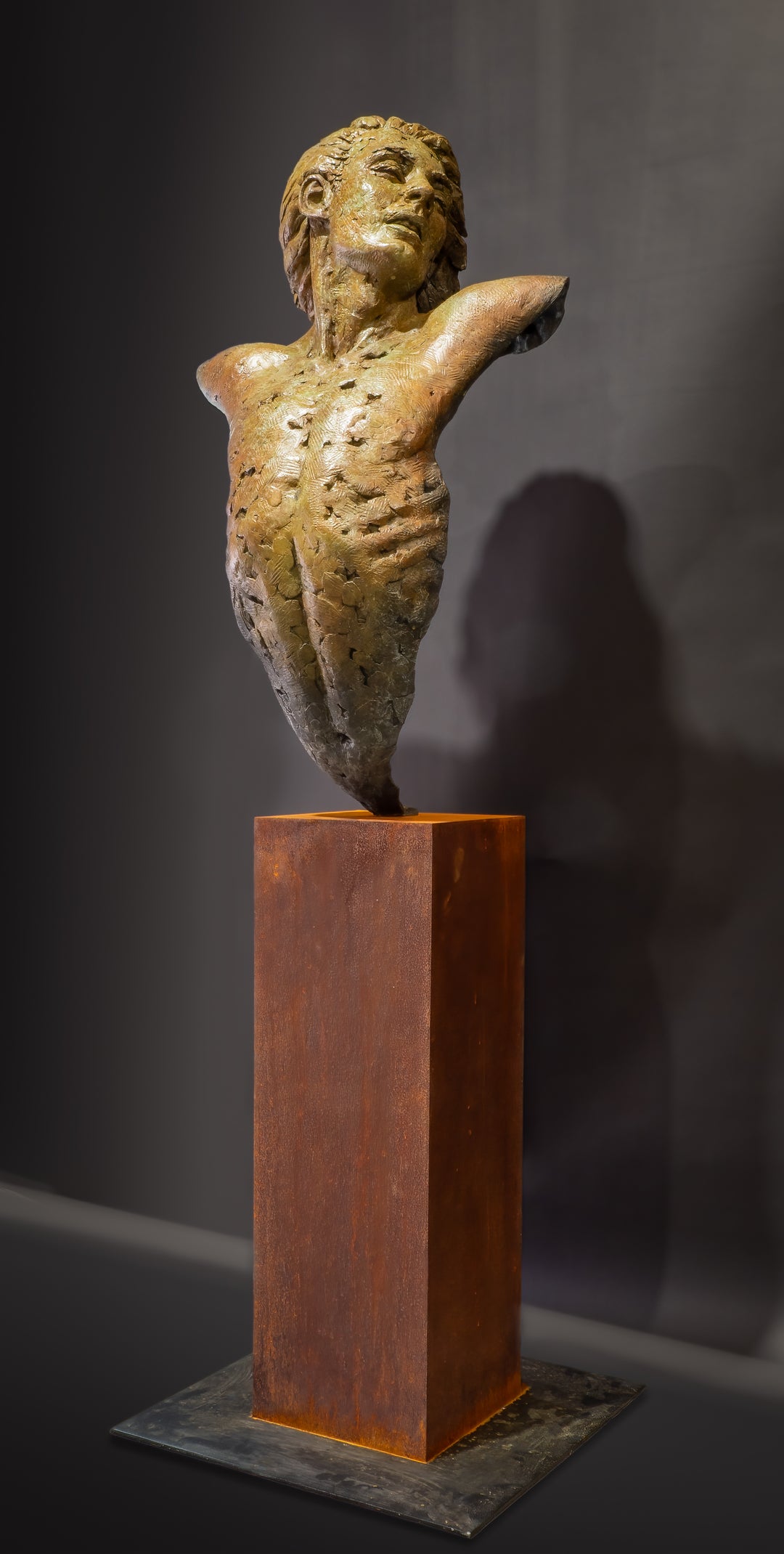 Nicola Godden Bliss Icarus bronze sculpture portraying a stylised human figure rising upward with arms extended and head turned, evoking a moment of release and quiet strength. The elongated form suggests lift and motion while remaining grounded through a solid Corten steel base. The textured bronze surface carries depth and variation, with light catching across its contours to enhance the sense of movement and presence.