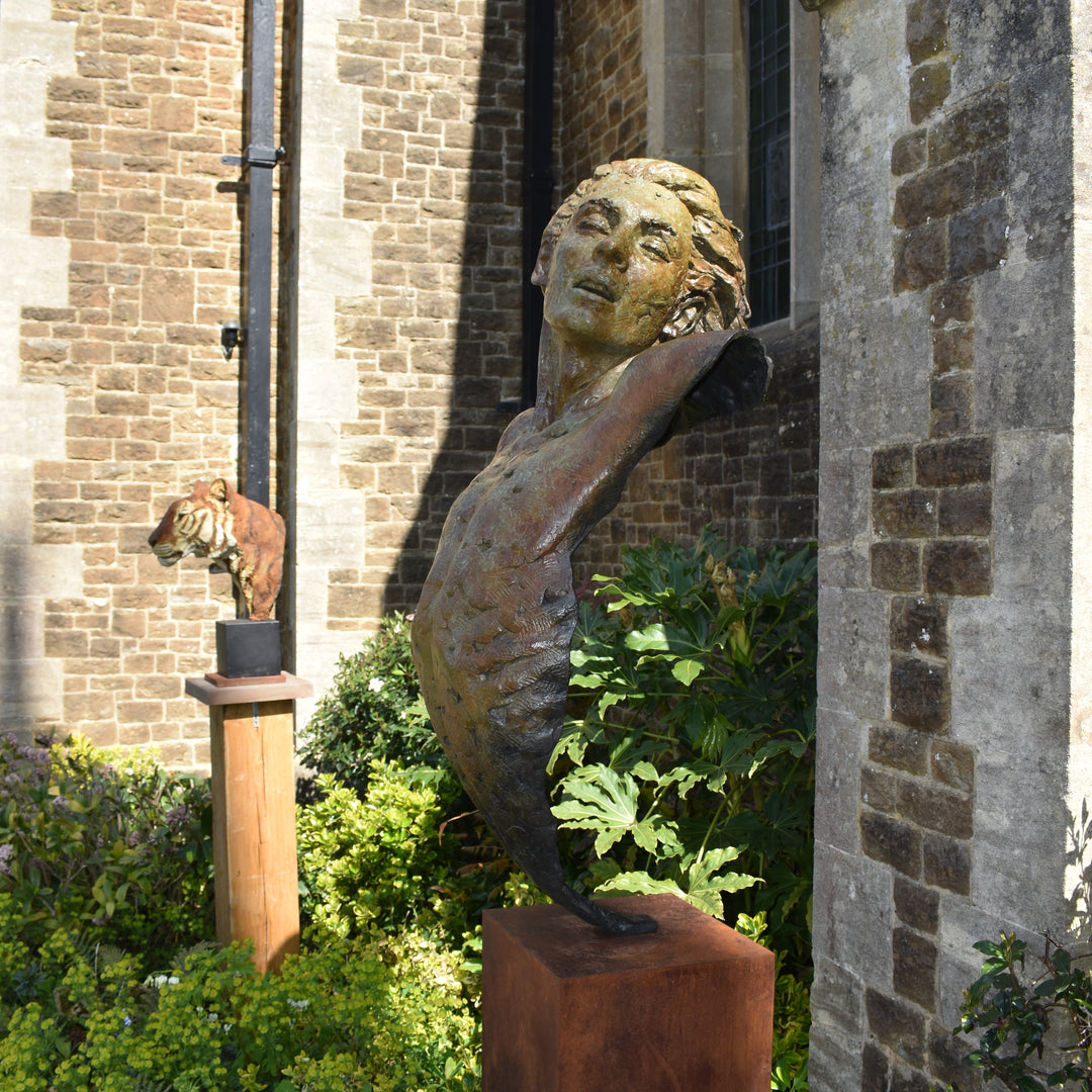 Nicola Godden Bliss Icarus bronze sculpture portraying a stylised human figure rising upward with arms extended and head turned, evoking a moment of release and quiet strength. The elongated form suggests lift and motion while remaining grounded through a solid Corten steel base. The textured bronze surface carries depth and variation, with light catching across its contours to enhance the sense of movement and presence.