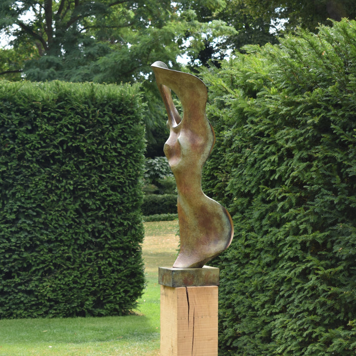 Abstract bronze sculpture of a stylised female torso with flowing curves and a hollowed circular form near the head, displayed in various outdoor and indoor settings. The sculpture has a rich patinated surface with green, gold, and bronze tones.