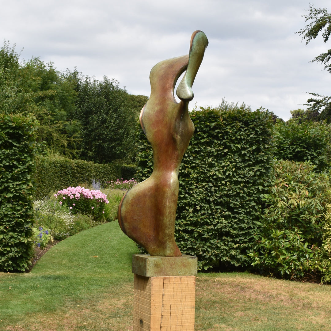 Abstract bronze sculpture of a stylised female torso with flowing curves and a hollowed circular form near the head, displayed in various outdoor and indoor settings. The sculpture has a rich patinated surface with green, gold, and bronze tones.