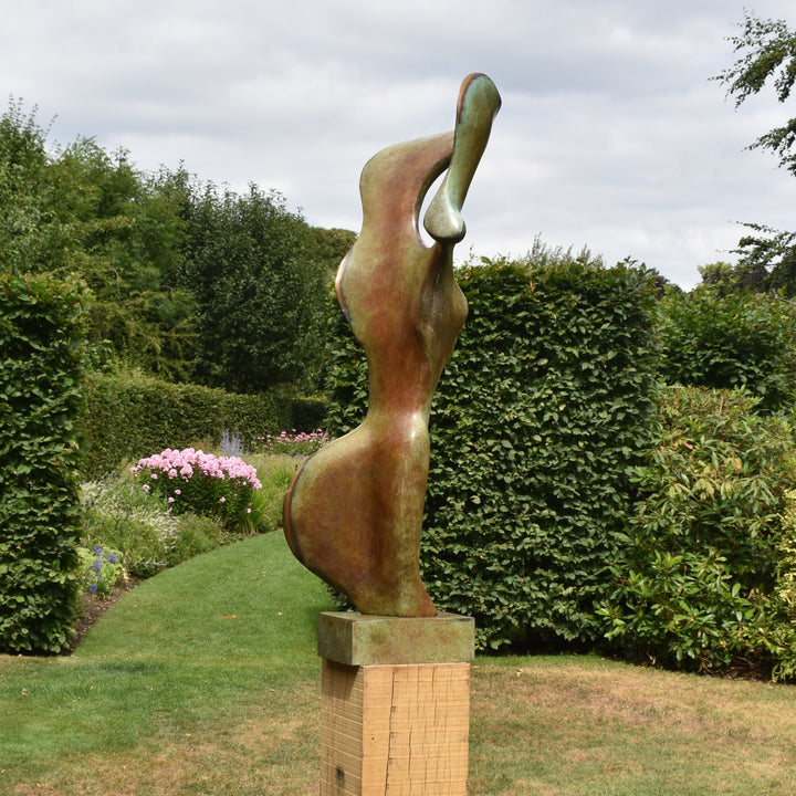 Abstract bronze sculpture of a stylised female torso with flowing curves and a hollowed circular form near the head, displayed in various outdoor and indoor settings. The sculpture has a rich patinated surface with green, gold, and bronze tones.