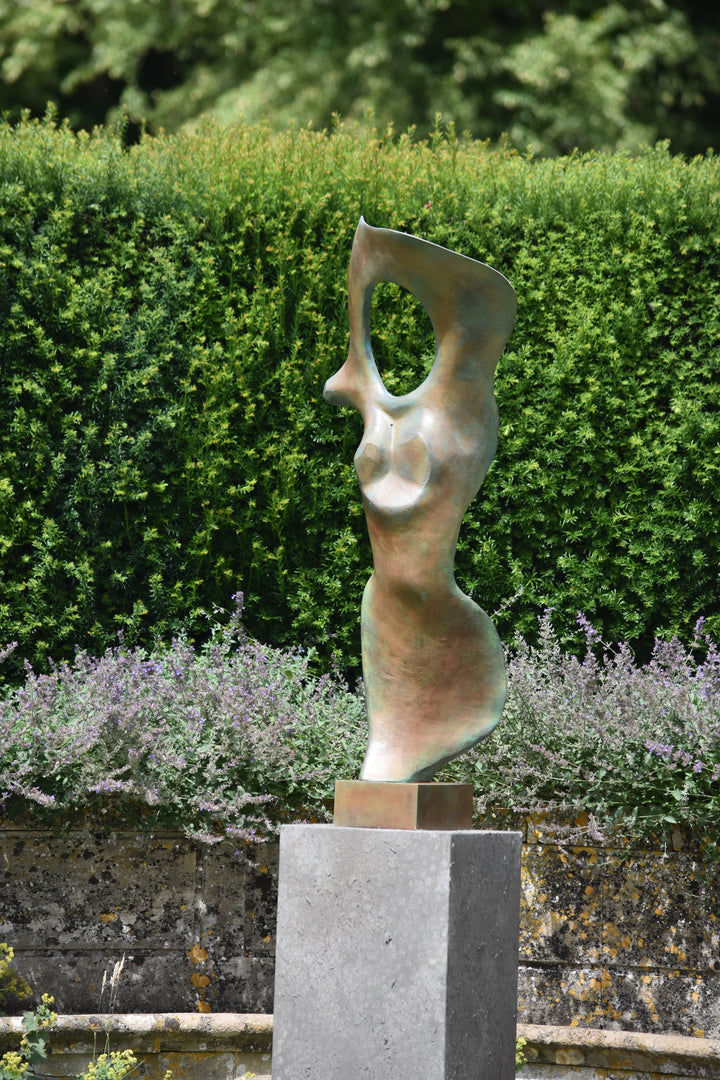 Abstract bronze sculpture of a stylised female torso with flowing curves and a hollowed circular form near the head, displayed in various outdoor and indoor settings. The sculpture has a rich patinated surface with green, gold, and bronze tones.
