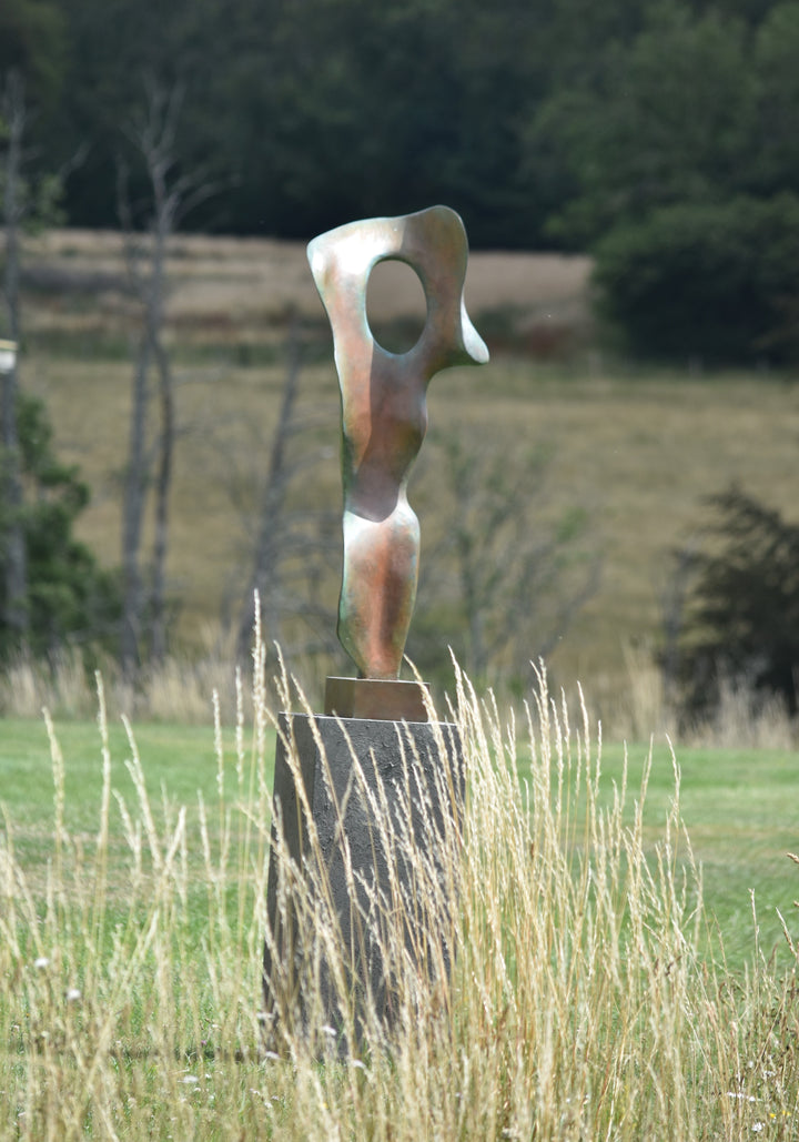 Abstract bronze sculpture of a stylised female torso with flowing curves and a hollowed circular form near the head, displayed in various outdoor and indoor settings. The sculpture has a rich patinated surface with green, gold, and bronze tones.