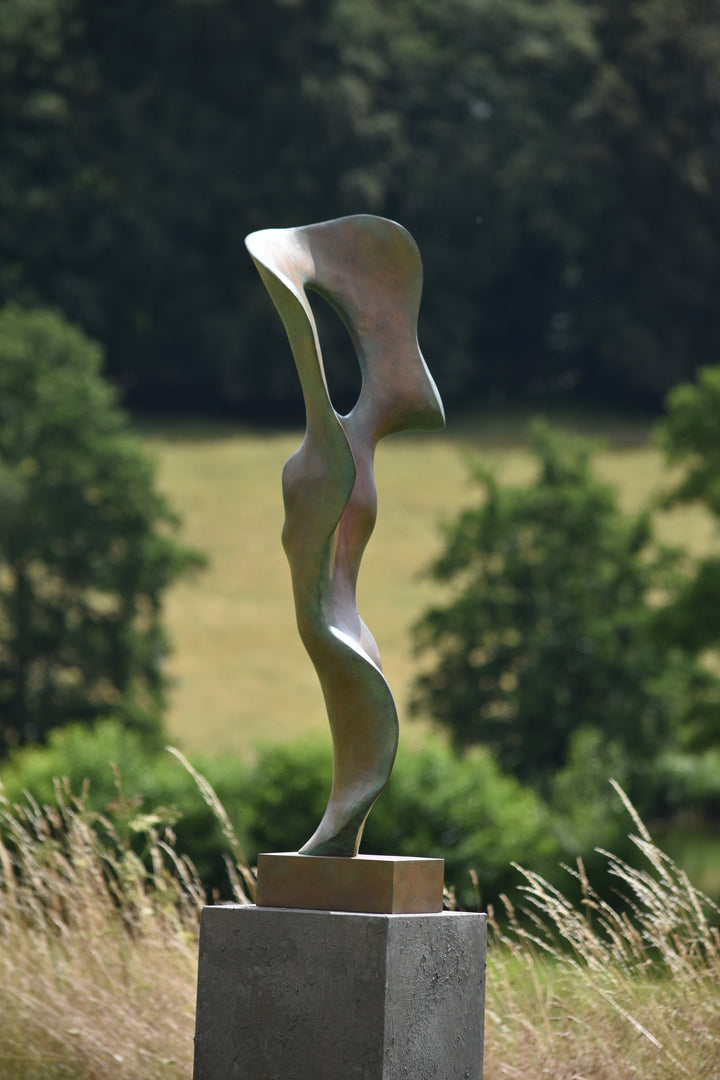 Abstract bronze sculpture of a stylised female torso with flowing curves and a hollowed circular form near the head, displayed in various outdoor and indoor settings. The sculpture has a rich patinated surface with green, gold, and bronze tones.