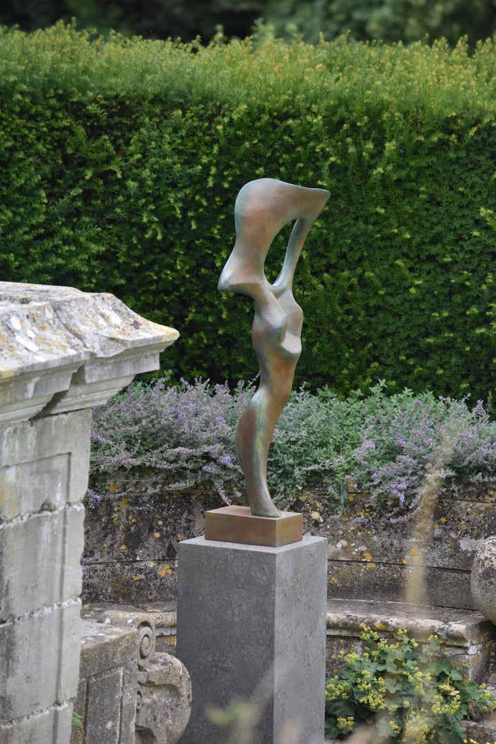 Abstract bronze sculpture of a stylised female torso with flowing curves and a hollowed circular form near the head, displayed in various outdoor and indoor settings. The sculpture has a rich patinated surface with green, gold, and bronze tones.