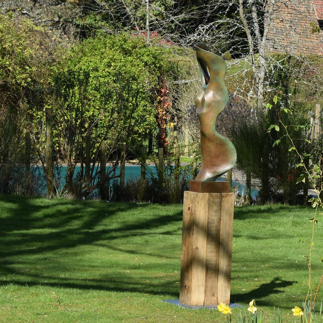 Abstract bronze sculpture of a stylised female torso with flowing curves and a hollowed circular form near the head, displayed in various outdoor and indoor settings. The sculpture has a rich patinated surface with green, gold, and bronze tones.