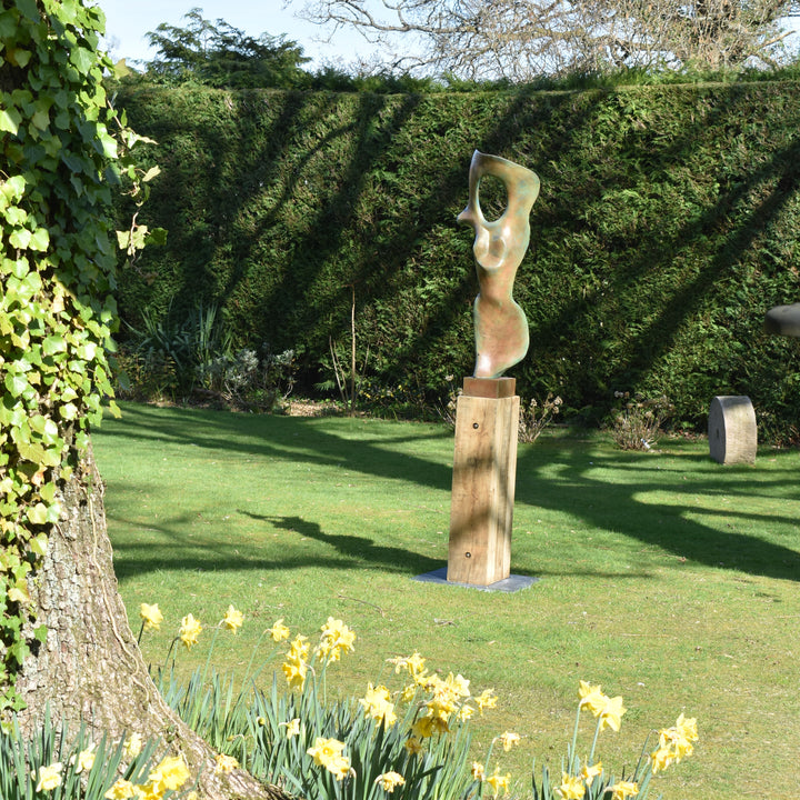 Abstract bronze sculpture of a stylised female torso with flowing curves and a hollowed circular form near the head, displayed in various outdoor and indoor settings. The sculpture has a rich patinated surface with green, gold, and bronze tones.