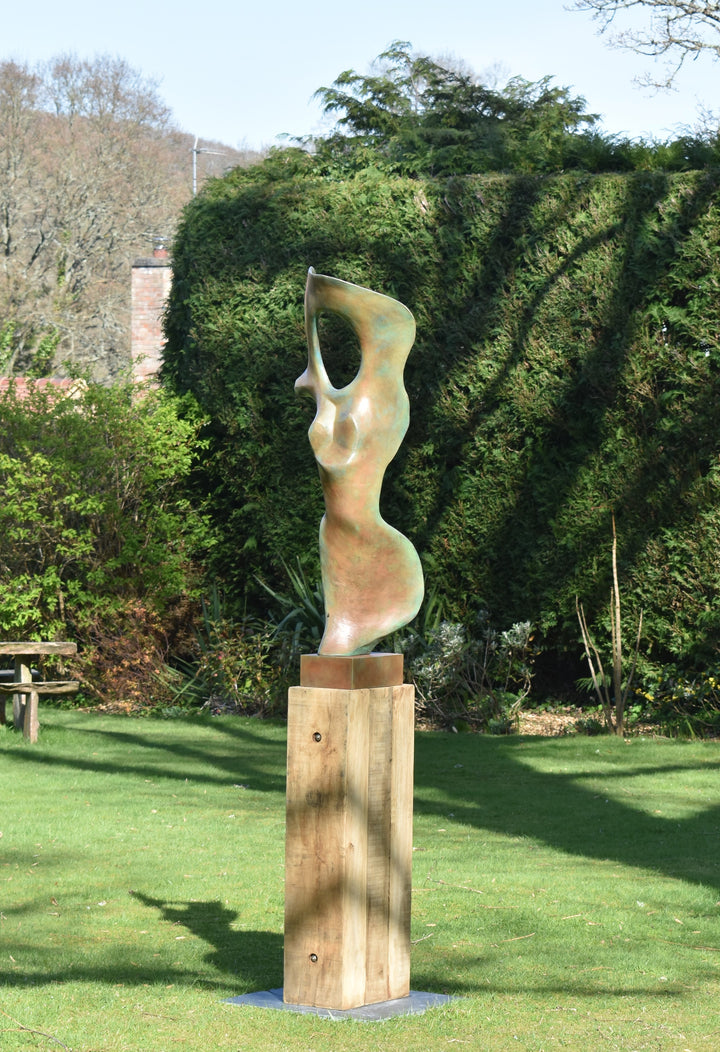 Abstract bronze sculpture of a stylised female torso with flowing curves and a hollowed circular form near the head, displayed in various outdoor and indoor settings. The sculpture has a rich patinated surface with green, gold, and bronze tones.
