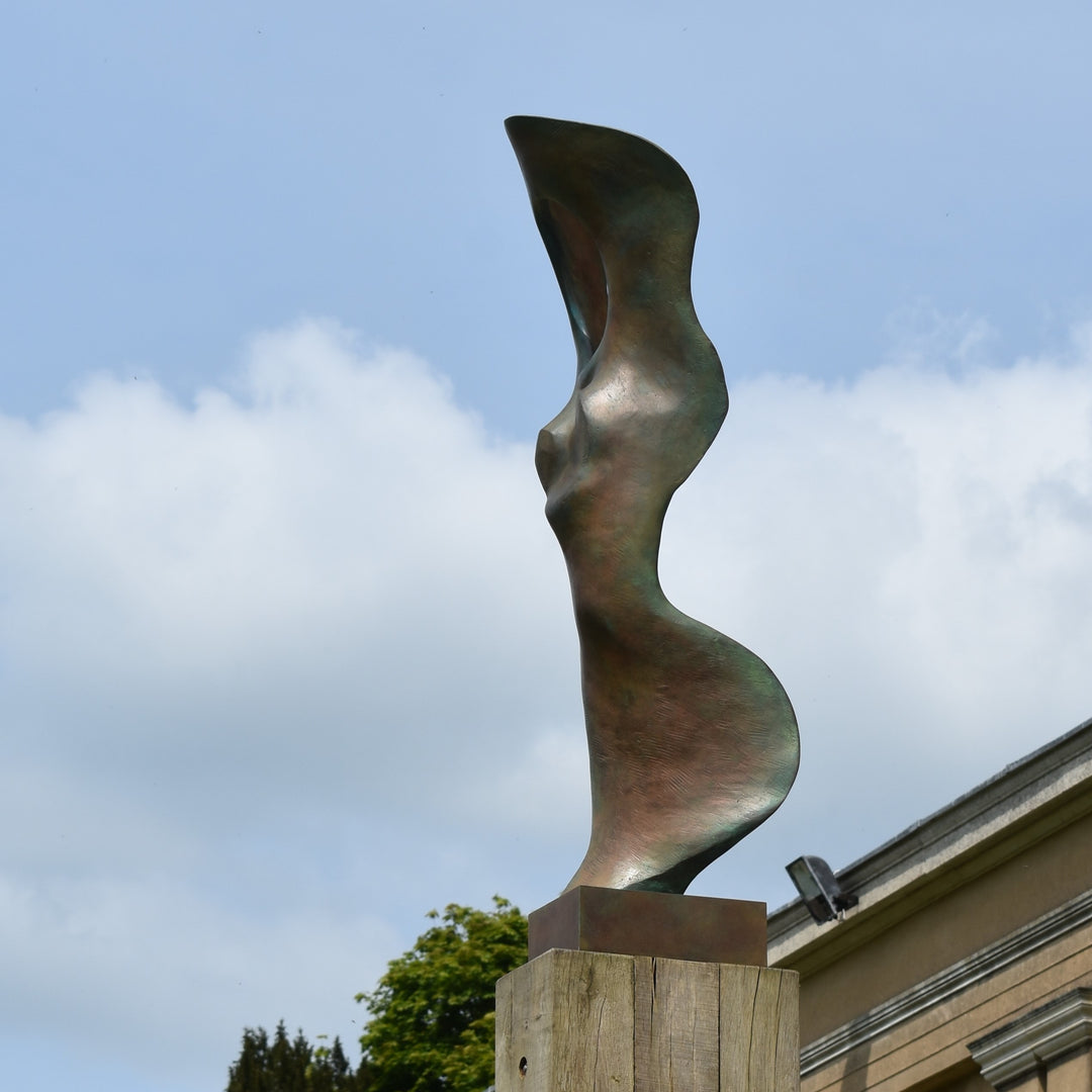 Abstract bronze sculpture of a stylised female torso with flowing curves and a hollowed circular form near the head, displayed in various outdoor and indoor settings. The sculpture has a rich patinated surface with green, gold, and bronze tones.