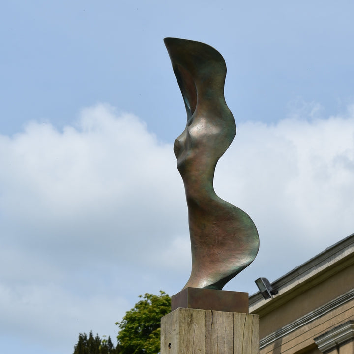 Abstract bronze sculpture of a stylised female torso with flowing curves and a hollowed circular form near the head, displayed in various outdoor and indoor settings. The sculpture has a rich patinated surface with green, gold, and bronze tones.