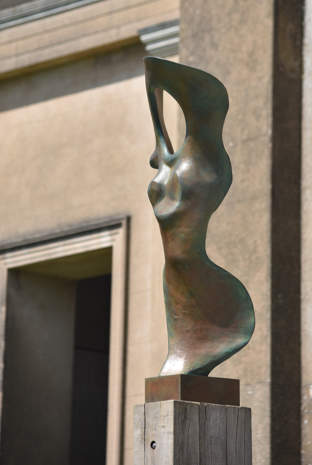 Abstract bronze sculpture of a stylised female torso with flowing curves and a hollowed circular form near the head, displayed in various outdoor and indoor settings. The sculpture has a rich patinated surface with green, gold, and bronze tones.