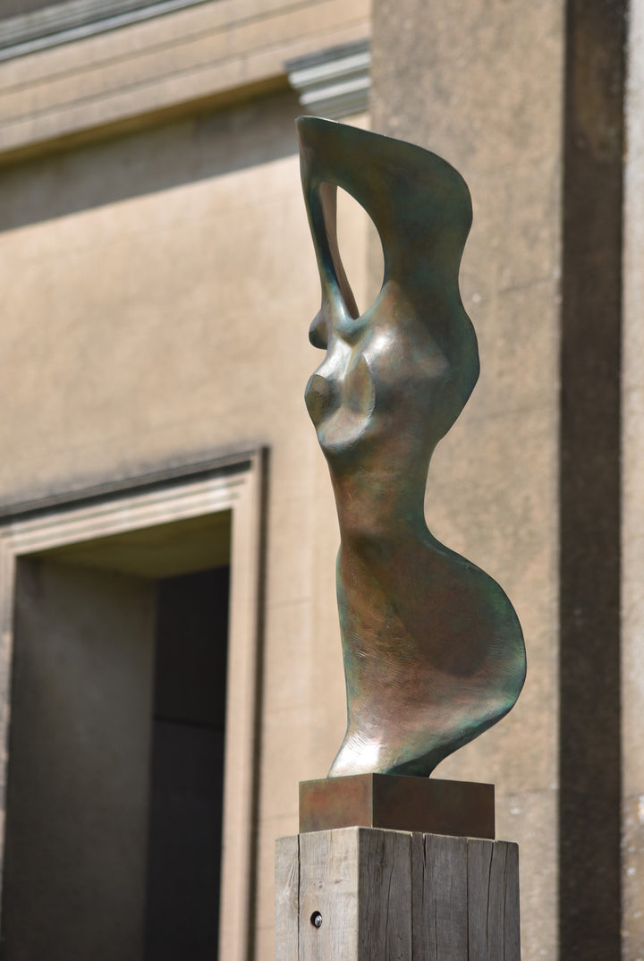 Abstract bronze sculpture of a stylised female torso with flowing curves and a hollowed circular form near the head, displayed in various outdoor and indoor settings. The sculpture has a rich patinated surface with green, gold, and bronze tones.