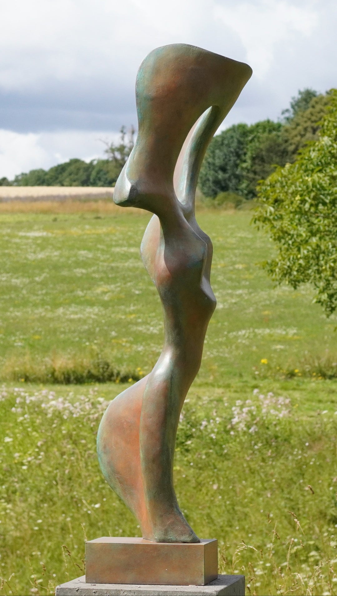 Abstract bronze sculpture of a stylised female torso with flowing curves and a hollowed circular form near the head, displayed in various outdoor and indoor settings. The sculpture has a rich patinated surface with green, gold, and bronze tones.