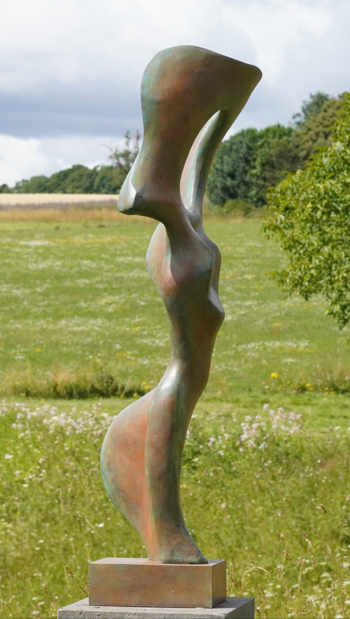 Abstract bronze sculpture of a stylised female torso with flowing curves and a hollowed circular form near the head, displayed in various outdoor and indoor settings. The sculpture has a rich patinated surface with green, gold, and bronze tones.