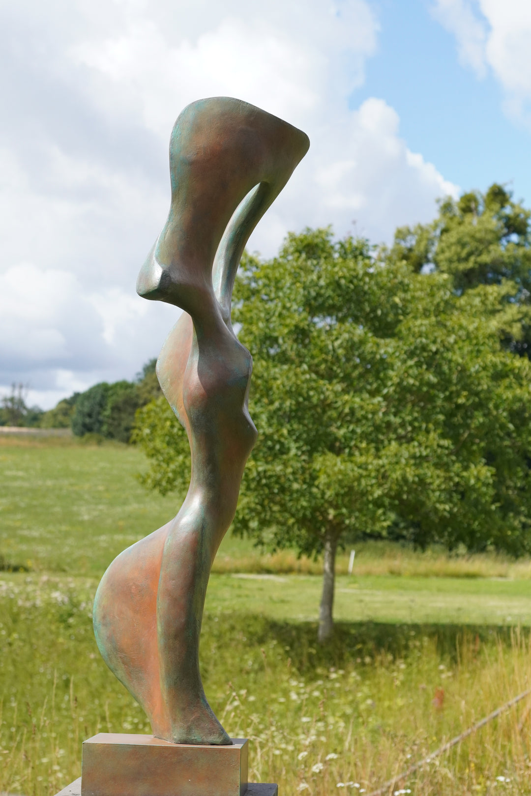 Abstract bronze sculpture of a stylised female torso with flowing curves and a hollowed circular form near the head, displayed in various outdoor and indoor settings. The sculpture has a rich patinated surface with green, gold, and bronze tones.