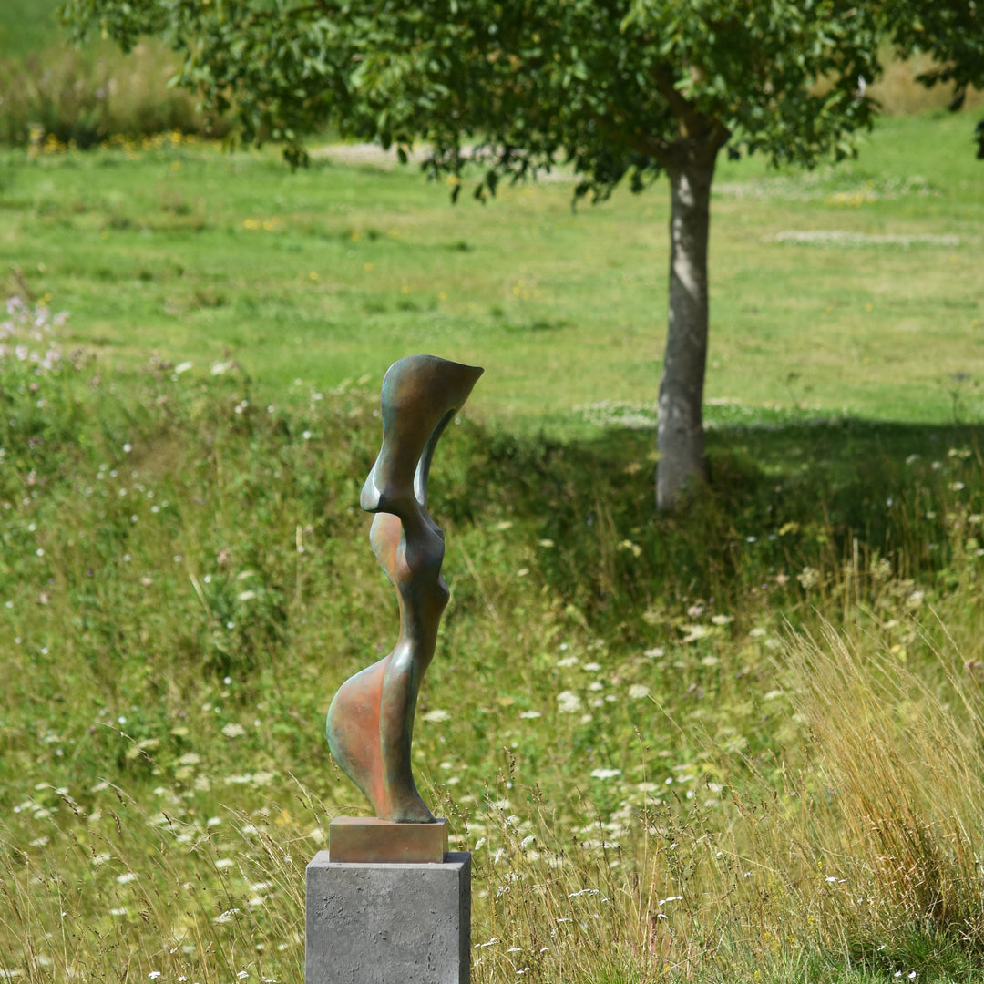Abstract bronze sculpture of a stylised female torso with flowing curves and a hollowed circular form near the head, displayed in various outdoor and indoor settings. The sculpture has a rich patinated surface with green, gold, and bronze tones.