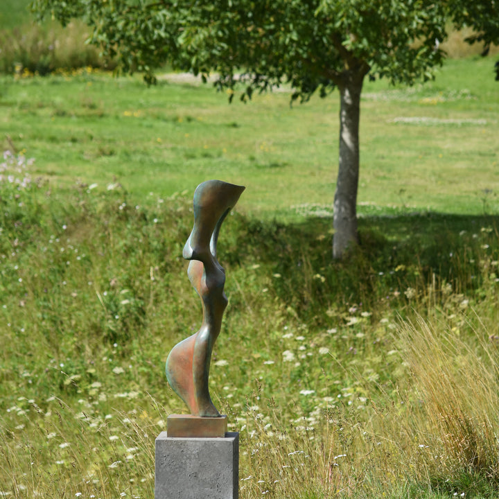 Abstract bronze sculpture of a stylised female torso with flowing curves and a hollowed circular form near the head, displayed in various outdoor and indoor settings. The sculpture has a rich patinated surface with green, gold, and bronze tones.