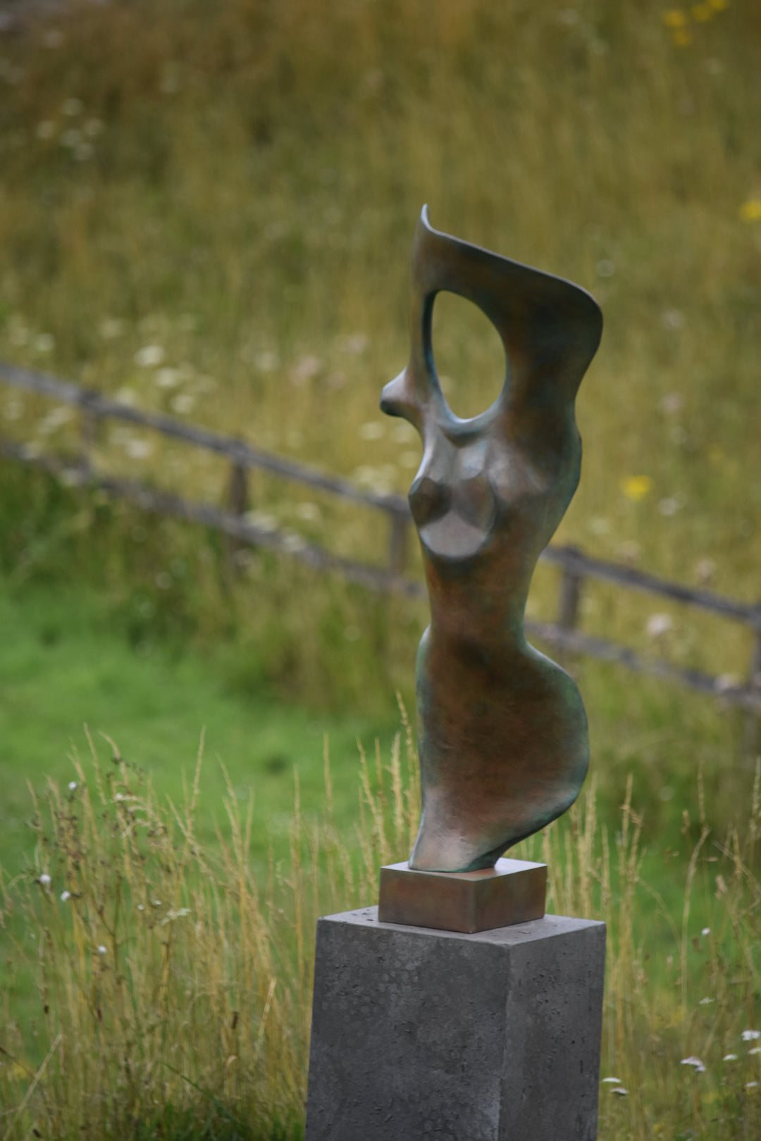 Abstract bronze sculpture of a stylised female torso with flowing curves and a hollowed circular form near the head, displayed in various outdoor and indoor settings. The sculpture has a rich patinated surface with green, gold, and bronze tones.