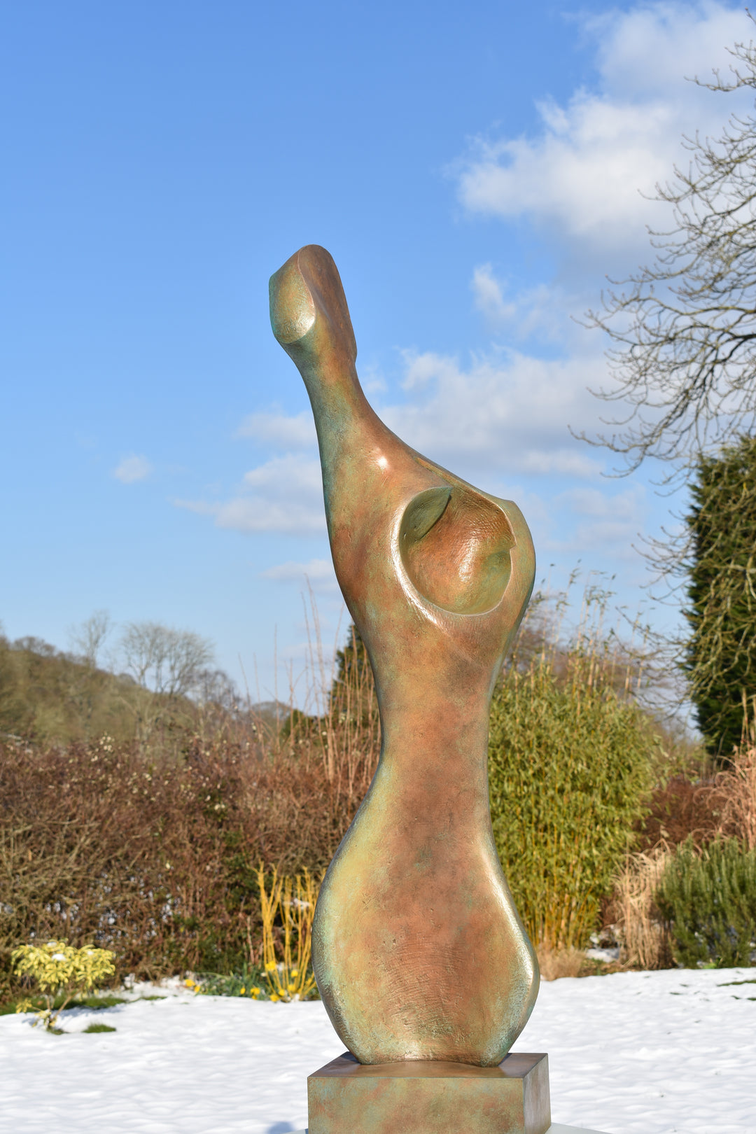 Bone Form IV — abstract bronze sculpture by Nicola Godden inspired by the natural curves and hollows of animal bone. The piece has a tall, sinuous form with a hollowed centre, organic surface texture and warm bronze patina shifting from earthy brown to soft green and gold highlights. Displayed outdoors on a one-metre timber plinth against a bright blue sky and winter landscape.