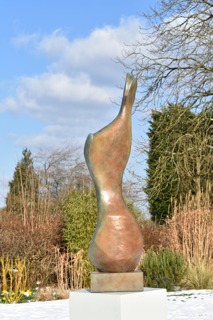 Bone Form IV — abstract bronze sculpture by Nicola Godden inspired by the natural curves and hollows of animal bone. The piece has a tall, sinuous form with a hollowed centre, organic surface texture and warm bronze patina shifting from earthy brown to soft green and gold highlights. Displayed outdoors on a one-metre timber plinth against a bright blue sky and winter landscape.