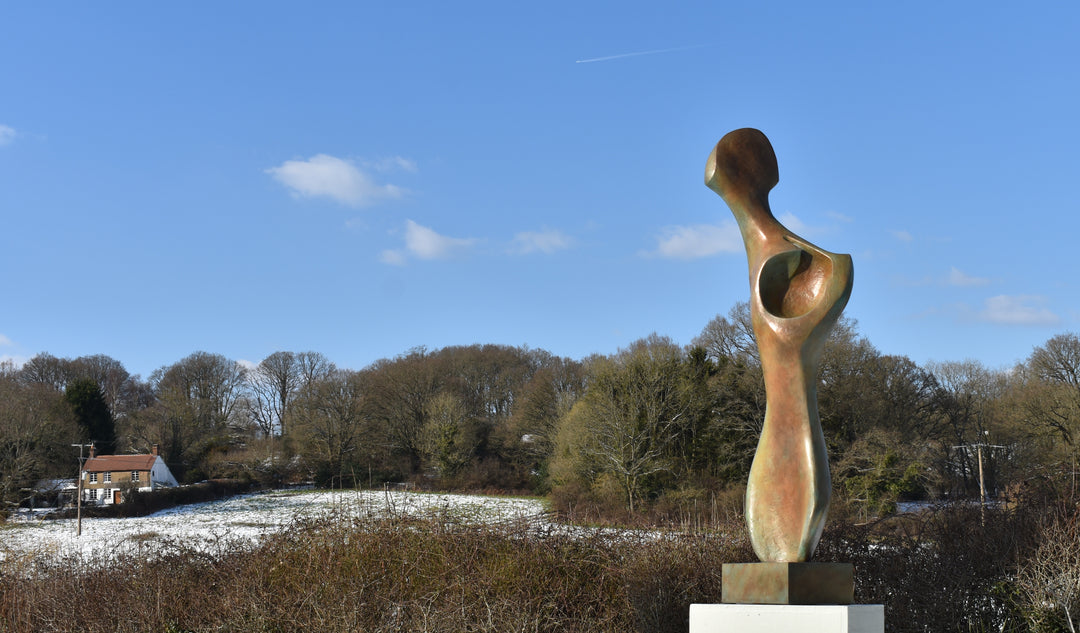 Bone Form IV — abstract bronze sculpture by Nicola Godden inspired by the natural curves and hollows of animal bone. The piece has a tall, sinuous form with a hollowed centre, organic surface texture and warm bronze patina shifting from earthy brown to soft green and gold highlights. Displayed outdoors on a one-metre timber plinth against a bright blue sky and winter landscape.