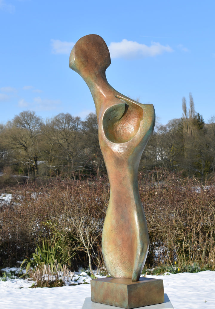 Bone Form IV — abstract bronze sculpture by Nicola Godden inspired by the natural curves and hollows of animal bone. The piece has a tall, sinuous form with a hollowed centre, organic surface texture and warm bronze patina shifting from earthy brown to soft green and gold highlights. Displayed outdoors on a one-metre timber plinth against a bright blue sky and winter landscape.