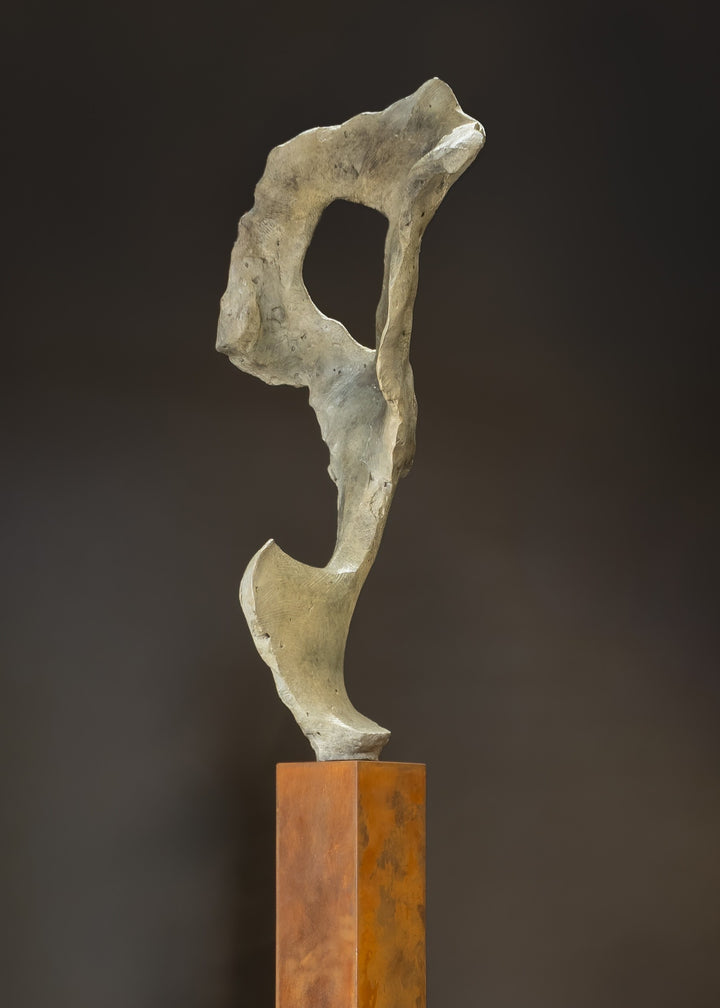 Abstract bronze sculpture by Nicola Godden titled “Bone Form VI”, featuring an elongated organic form with a hollowed negative space near the upper section, inspired by skeletal and geological structures. The sculpture rises vertically from a tall rusted steel plinth, with a softly textured, patinated bronze surface that emphasises erosion, balance, and the quiet strength of natural forms within contemporary sculptural practice.