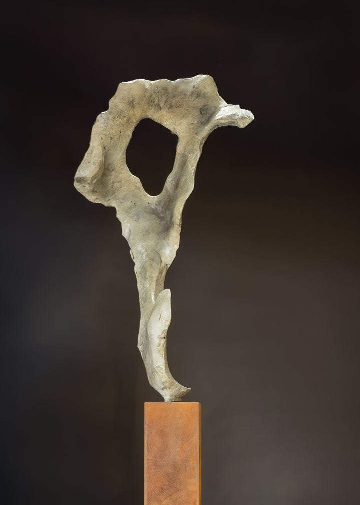 Abstract bronze sculpture by Nicola Godden titled “Bone Form VI”, featuring an elongated organic form with a hollowed negative space near the upper section, inspired by skeletal and geological structures. The sculpture rises vertically from a tall rusted steel plinth, with a softly textured, patinated bronze surface that emphasises erosion, balance, and the quiet strength of natural forms within contemporary sculptural practice.