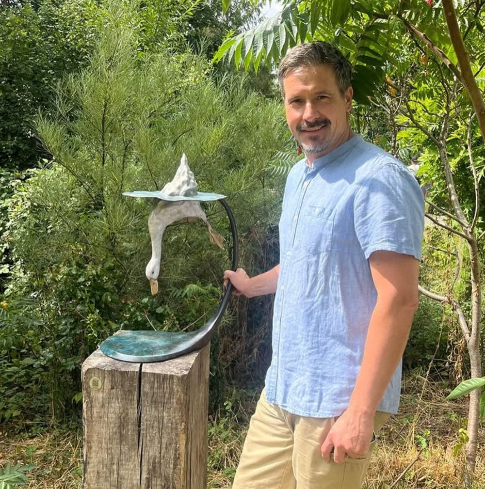 Playful bronze sculpture of a duck diving underwater, with only the rear and feet above a rippled surface, supported by a curved bronze stand — full of character and movement.
