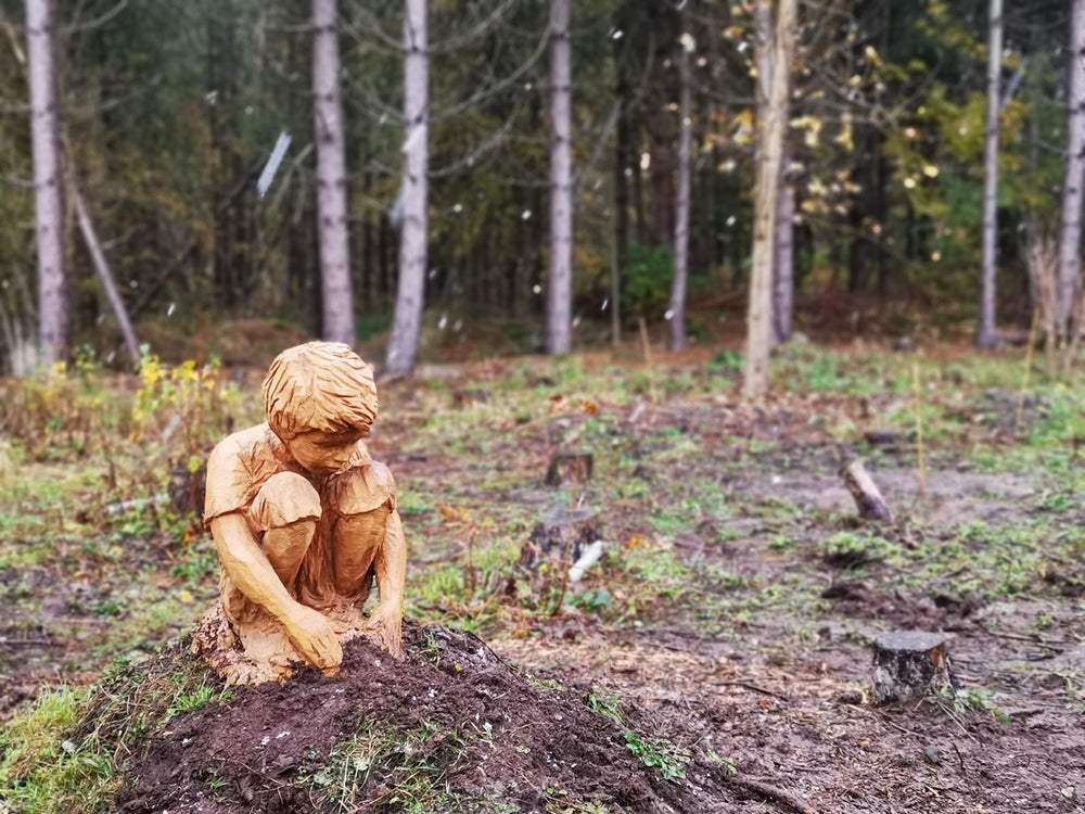 Hand-carved wooden sculpture of a young boy crouched down, intently focused as he digs or plays in the earth,  and plants a tree, set against a misty woodland backdrop.
