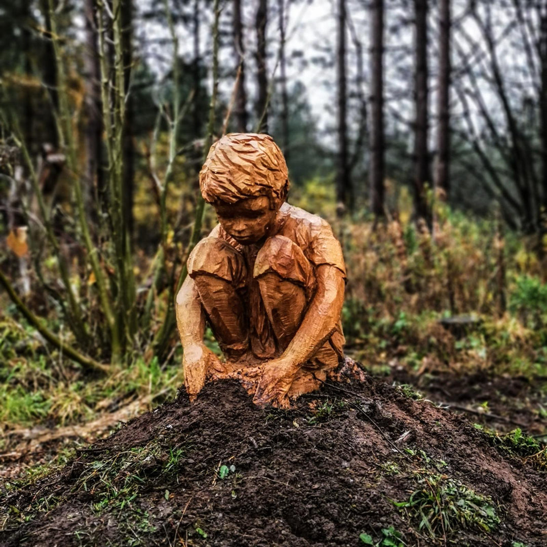 Hand-carved wooden sculpture of a young boy crouched down, intently focused as he digs or plays in the earth,  and plants a tree, set against a misty woodland backdrop.