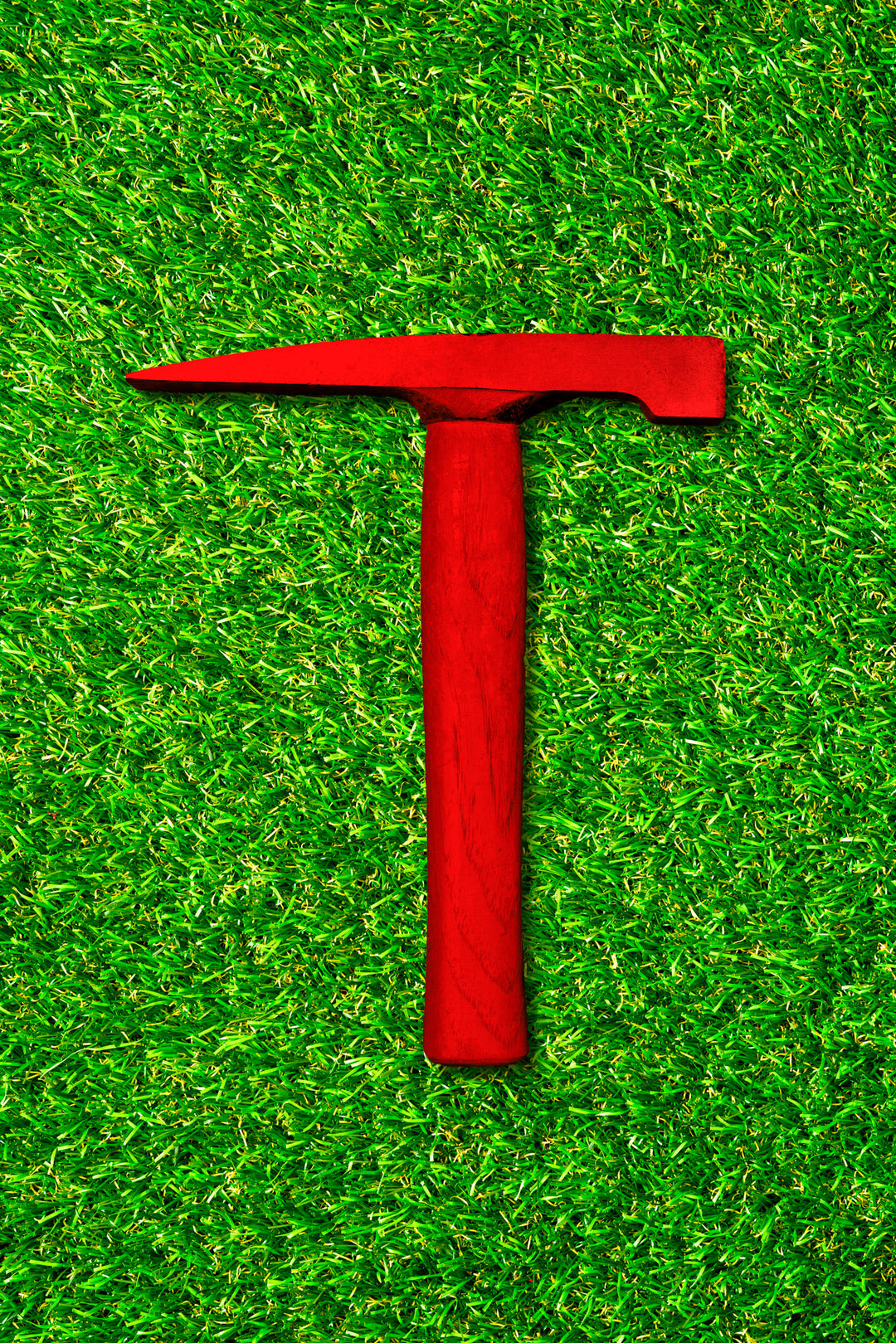 Photograph of a vivid red pick hammer placed vertically on a bright green grass background, creating a striking contrast in colour and form.