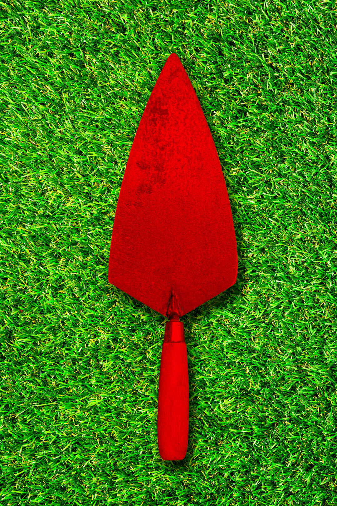 Photograph of a bright red pointing trowel placed upright on vivid green grass, creating a bold contrast between the tool and background.