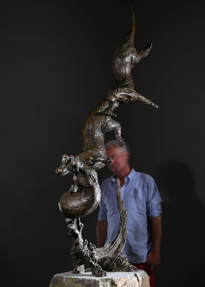 ‘Otters Playing Underwater’ by Hamish Mackie, a life-size limited-edition bronze wildlife sculpture depicting a mother otter and her playful cubs twisting, diving and swimming around a sculpted underwater tree root. The dynamic vertical composition features expressive animal movements, detailed fur textures and a weathered bronze finish in rich brown and earthy tones, capturing the playful energy and natural behaviour of otters beneath the water.
