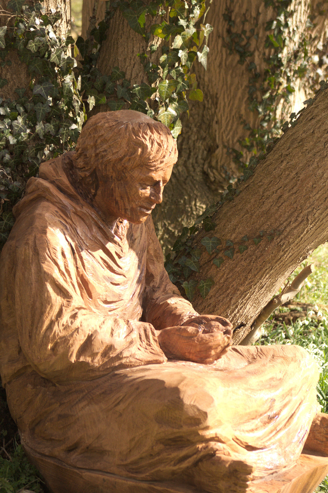 Wooden sculpture of a seated monk or robed figure carved in a naturalistic style, positioned against a tree trunk in a woodland setting. The figure gazes downward with a peaceful expression, cupping something gently in both hands. Ivy vines and bark textures surround the sculpture, enhancing its organic connection to the environment.