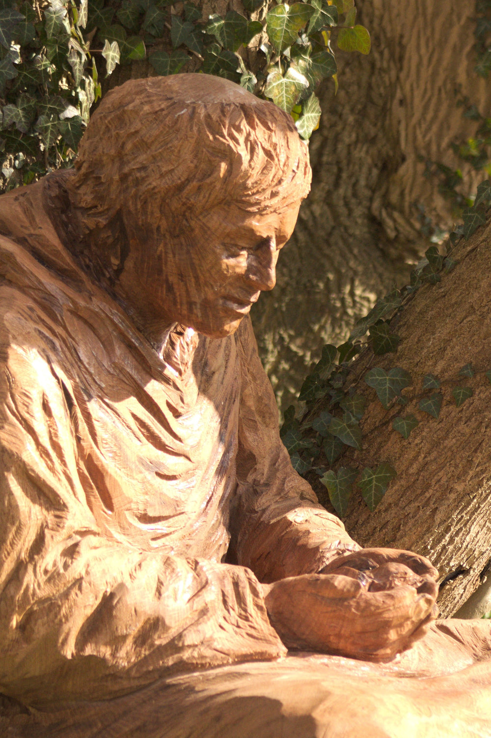 Wooden sculpture of a seated monk or robed figure carved in a naturalistic style, positioned against a tree trunk in a woodland setting. The figure gazes downward with a peaceful expression, cupping something gently in both hands. Ivy vines and bark textures surround the sculpture, enhancing its organic connection to the environment.
