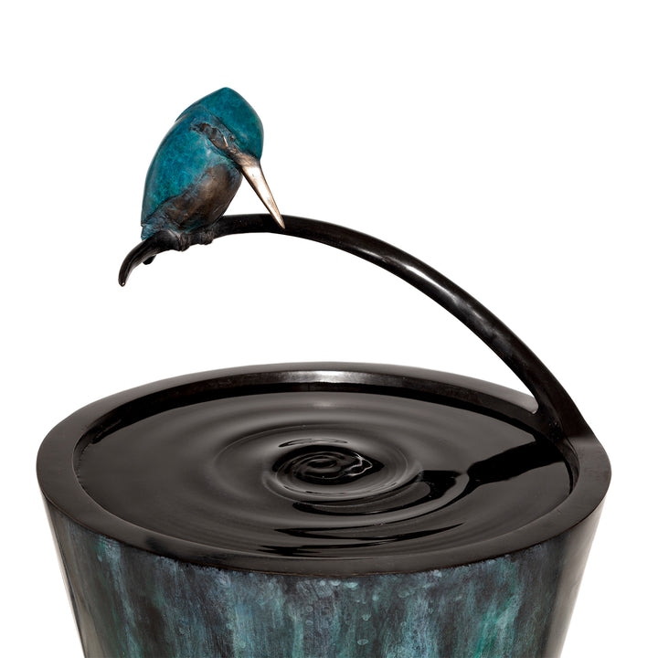 ‘Halcyon Days’ by Pat Bull, a limited-edition contemporary bronze wildlife sculpture and functional birdbath featuring a kingfisher perched on a curved bronze support above a circular basin designed to hold water. The bird features vivid turquoise and deep blue patina, a warm bronze breast and a polished golden beak, with its head lowered towards the reflective water below. The tall, tapered conical birdbath has a textured verdigris finish in charcoal, teal and blue-green tones, supported by a circular dark