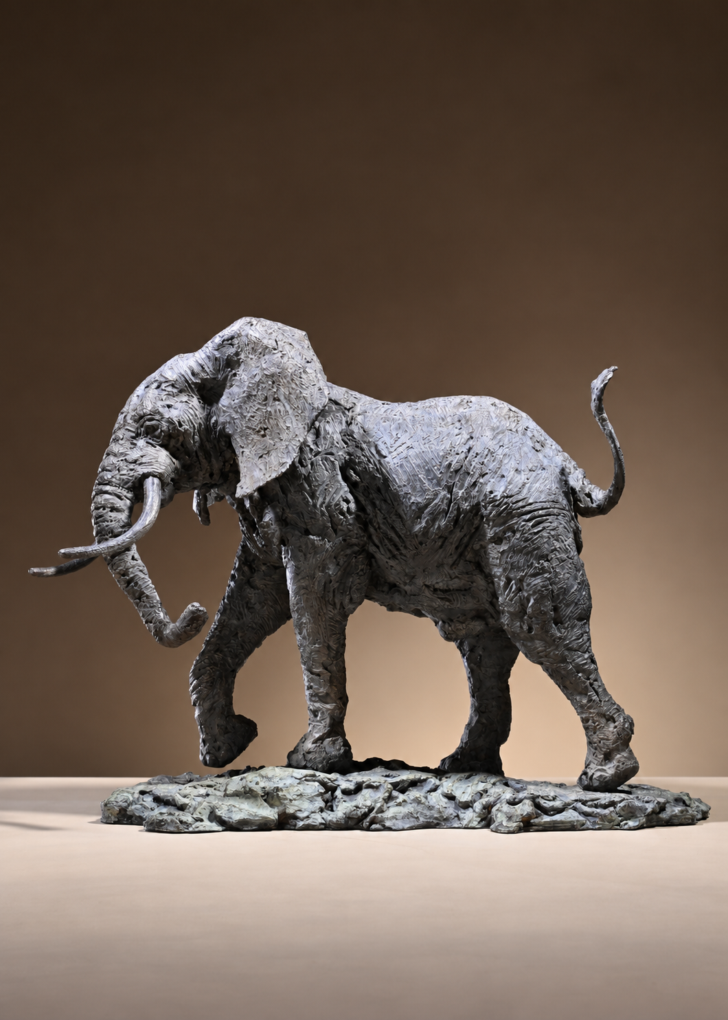 ‘Elephant Bull: Large’ by Hamish Mackie, a limited-edition bronze wildlife sculpture depicting a powerful African bull elephant stepping backwards with its head raised, trunk curled upwards, long curved tusks and tail lifted. Finished in dark grey bronze with richly textured skin and detailed muscular features, the sculpture captures the strength, intelligence and alert character of an African elephant standing on a rugged naturalistic base.