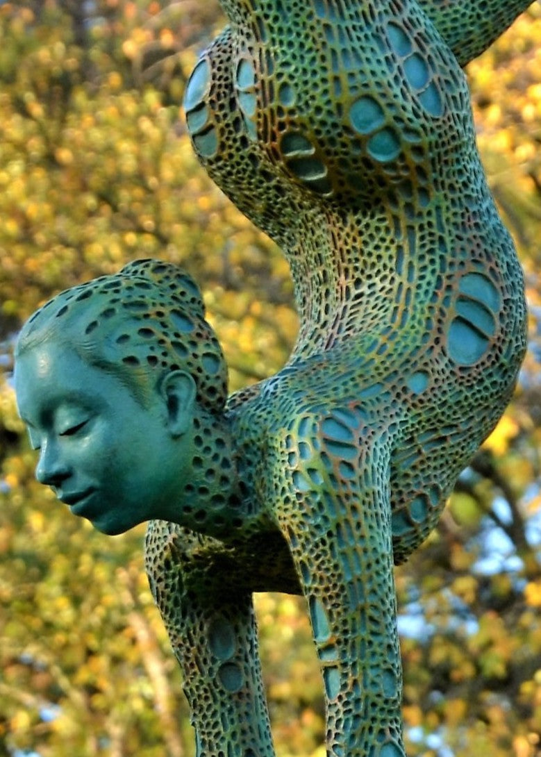 Bronze sculpture of a contorted female figure balanced in an acrobatic inverted pose, her body entirely covered in an intricate cellular pattern inspired by plant structures seen under a microscope. Painted in shades of aqua, teal, moss green and warm ochre, the sculpture feels alive with organic energy and movement. Set outdoors against soft greenery, the piece captures the idea of growth, vitality and the hidden structures of nature.