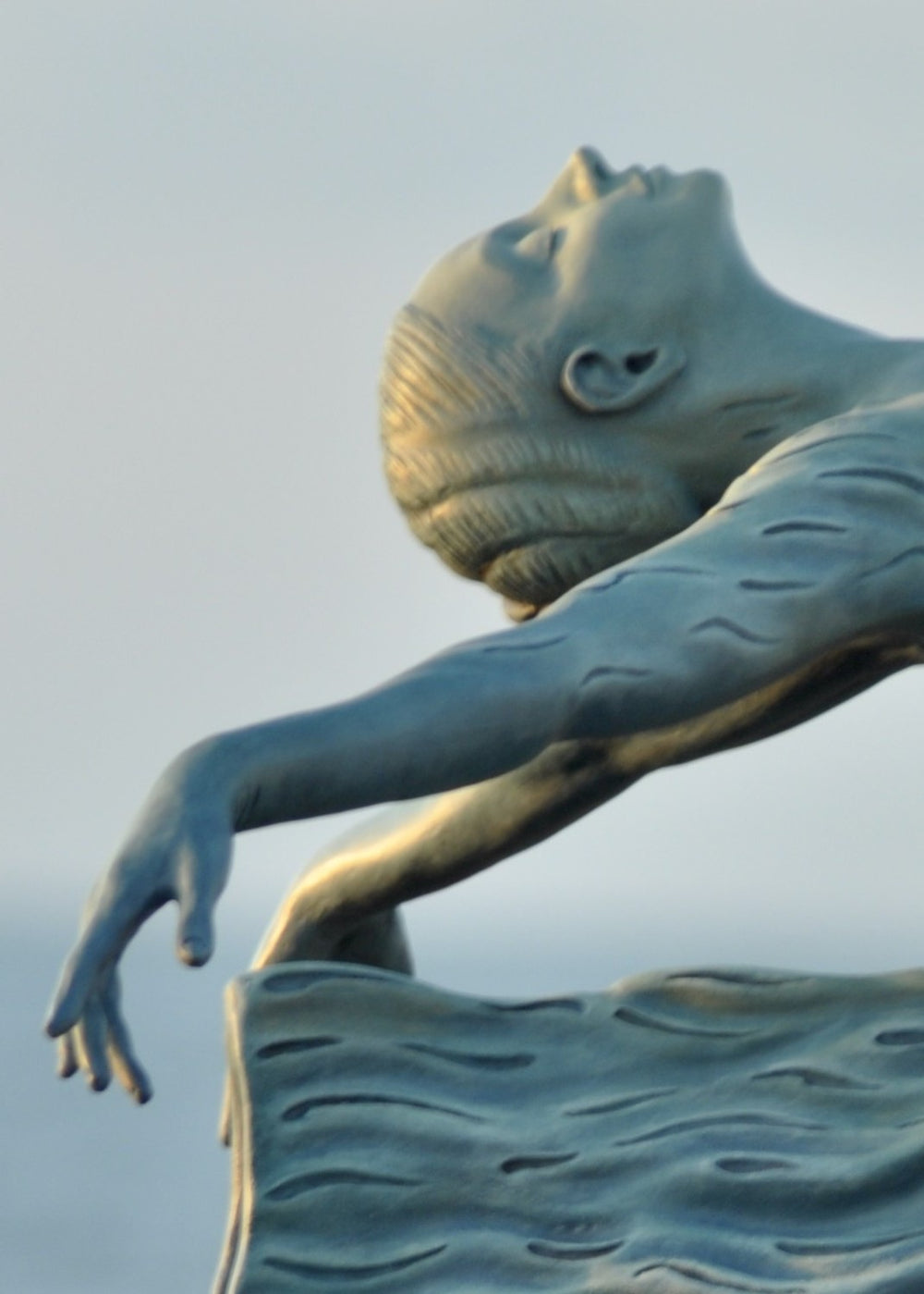 Bronze sculpture of a female figure leaning back in a flowing, wave-textured dress that echoes the motion of the sea, balanced delicately on one foot atop a plinth. Her arms and head are extended in a serene, open pose, with the sculpture photographed outdoors against a calm ocean horizon.