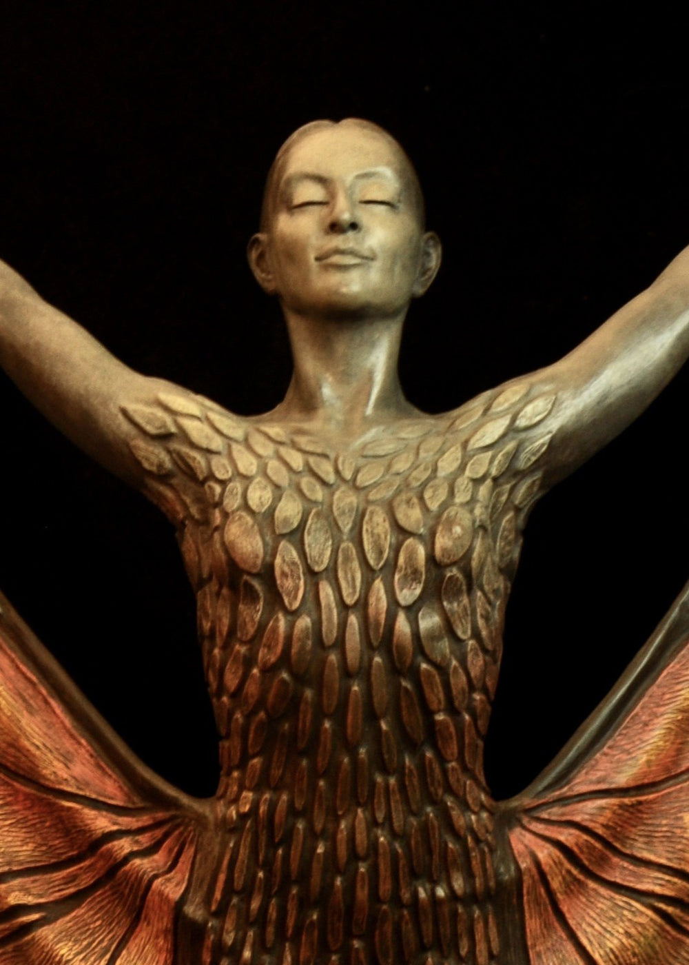 Bronze sculpture of a standing female figure lifting wide, wing-like panels of her dress, revealing intricate monarch butterfly patterns in warm orange, copper, gold and soft cream tones. Her expression is serene and confident, with arms raised and shoulders open as if taking flight. The textured surface and detailed markings mimic butterfly wings while the figure maintains a grounded, elegant posture. The sculpture conveys freedom, transformation and self-expression.