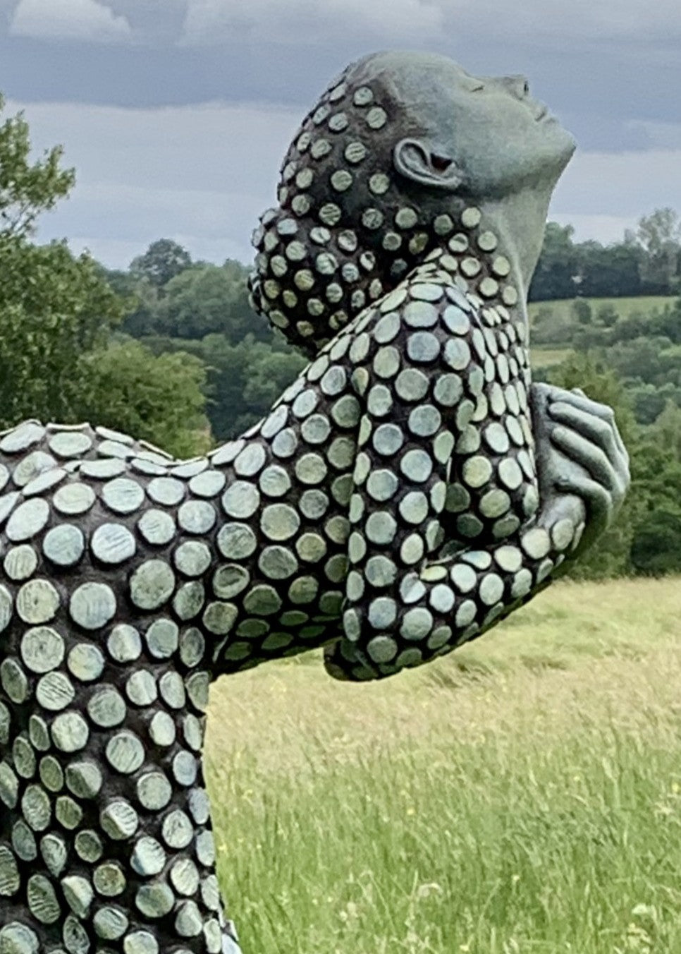 Bronze sculpture of a female figure poised on one leg atop a tiered plinth, her body arched back and arms drawn to her chest as she lifts her face toward the sky. Her dress and skin are patterned with circular, moon-like discs that flow outward into a crescent shape, photographed outdoors in a wide green landscape beneath a cloudy sky.