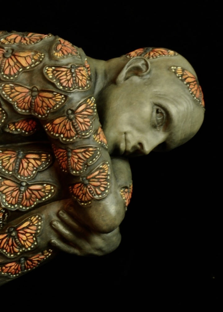 Bronze sculpture of a crouched male figure balanced on a square plinth, his body wrapped tightly around itself in a contemplative pose. The entire surface is patterned with vivid monarch butterflies in orange, black and soft yellow tones, creating a striking contrast against the subdued bronze of the skin. His expression is introspective and hesitant, poised between stillness and the potential for movement. Set against a dark background, the sculpture highlights themes of transformation and courage.