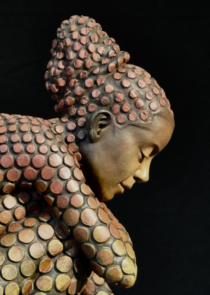 Bronze sculpture of a crouching female figure, her body covered in a gradual pattern of circular, sunset-toned discs that shift from warm reds to soft golds. She balances on a textured block, head bowed gently in a contemplative pose, with a dark backdrop emphasising the warmth and depth of the patina.